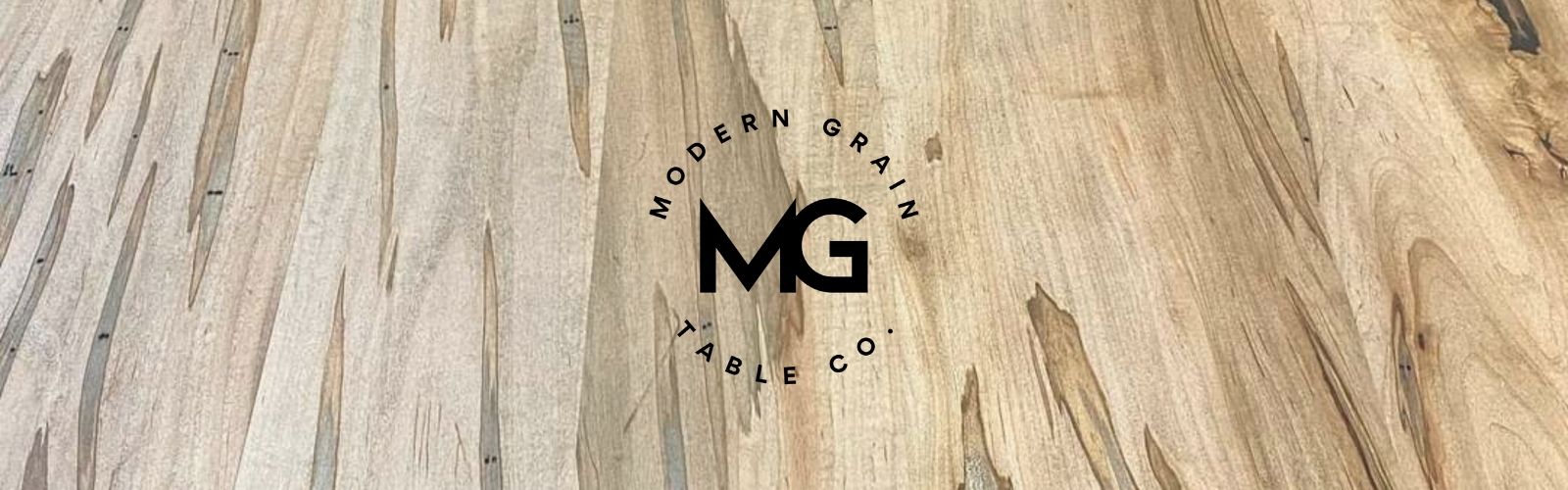 Modern Grain Table Co. logo over ambrosia maple hardwood grain – showcasing craftsmanship and premium custom table design