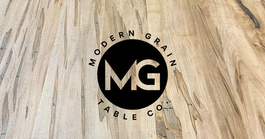 Modern Grain Table Co. logo over ambrosia maple hardwood grain background – handcrafted custom table brand based in Michigan